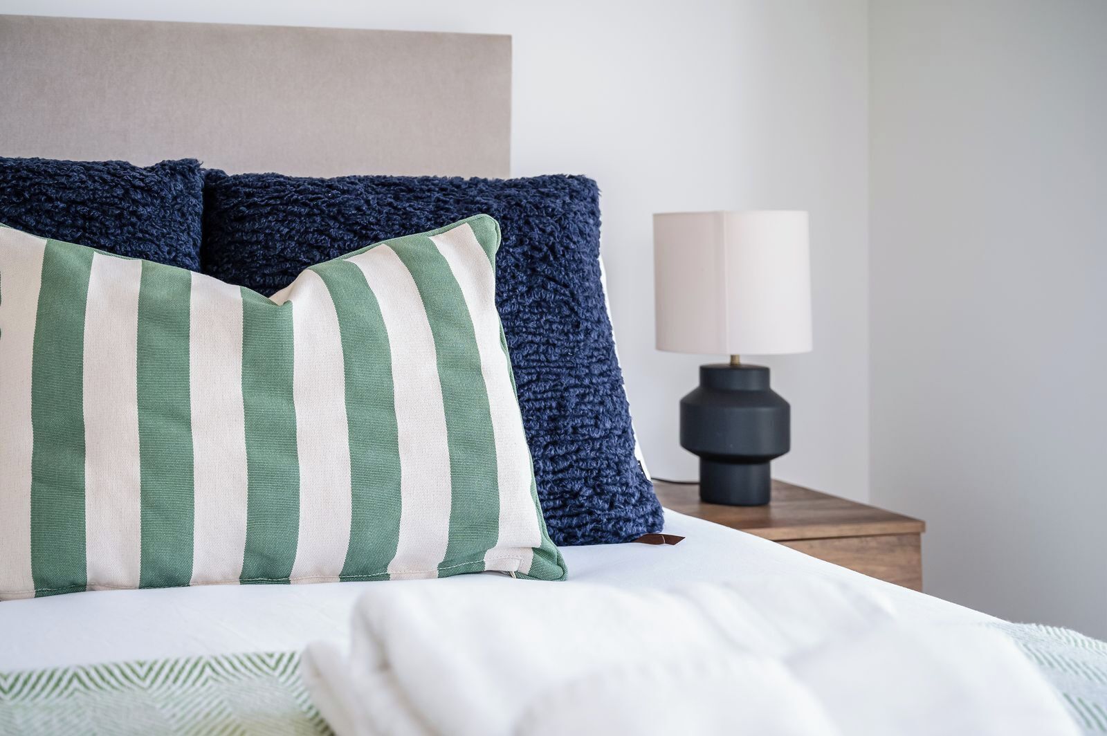 Una cama con almohadas a rayas, un cojín de textura oscura y una lámpara de noche sobre una mesita de madera.