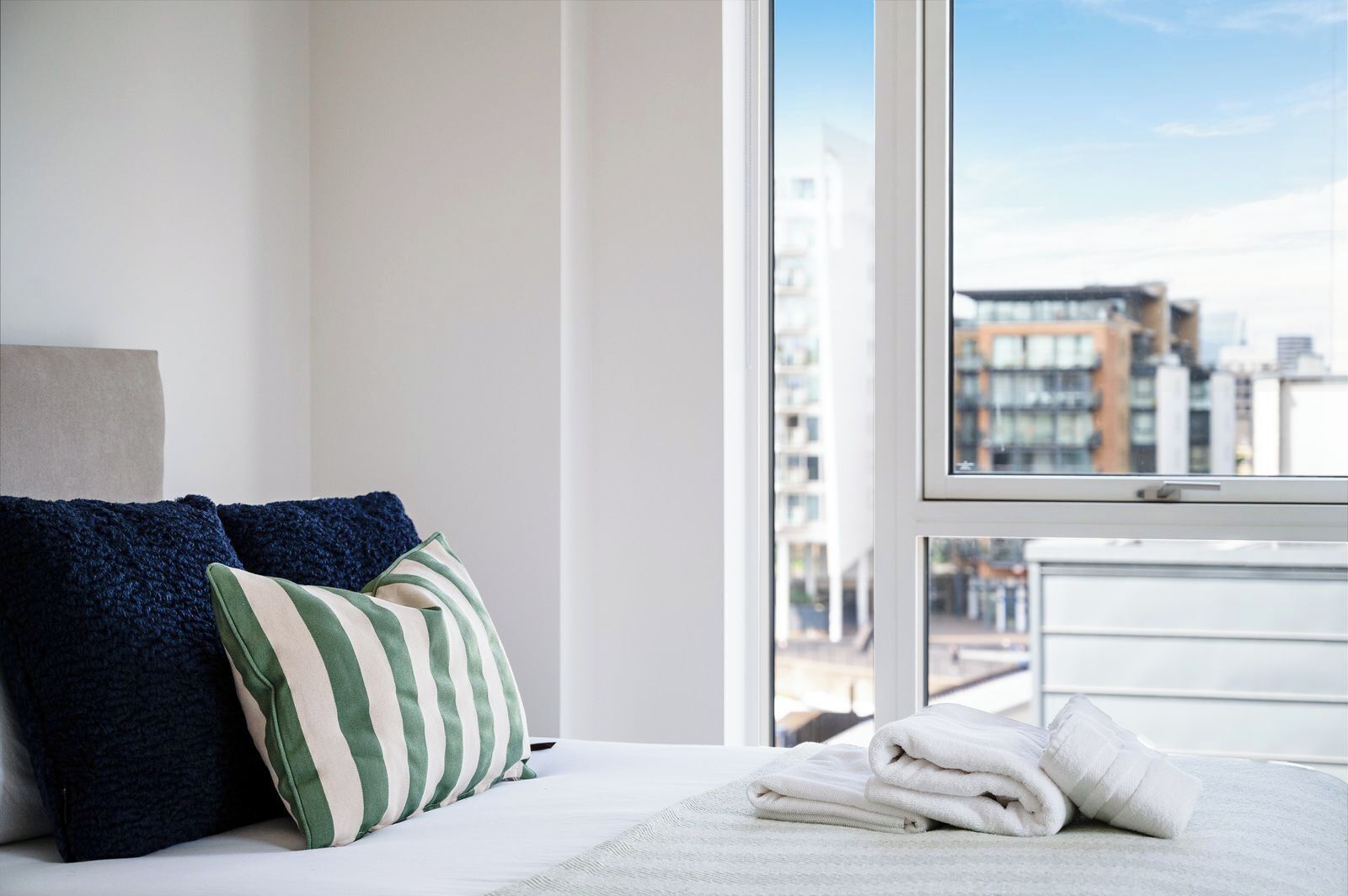 A hotel room with a bed, pillows, a window with a city view, and a neatly folded towel on the bed.