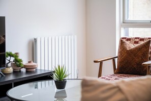A living room with a round glass table, a potted plant, a sofa with a patterned cushion, a television, and a radiator. - Elegant 2-Bed Retreat Near Canary Wharf (Greater London)