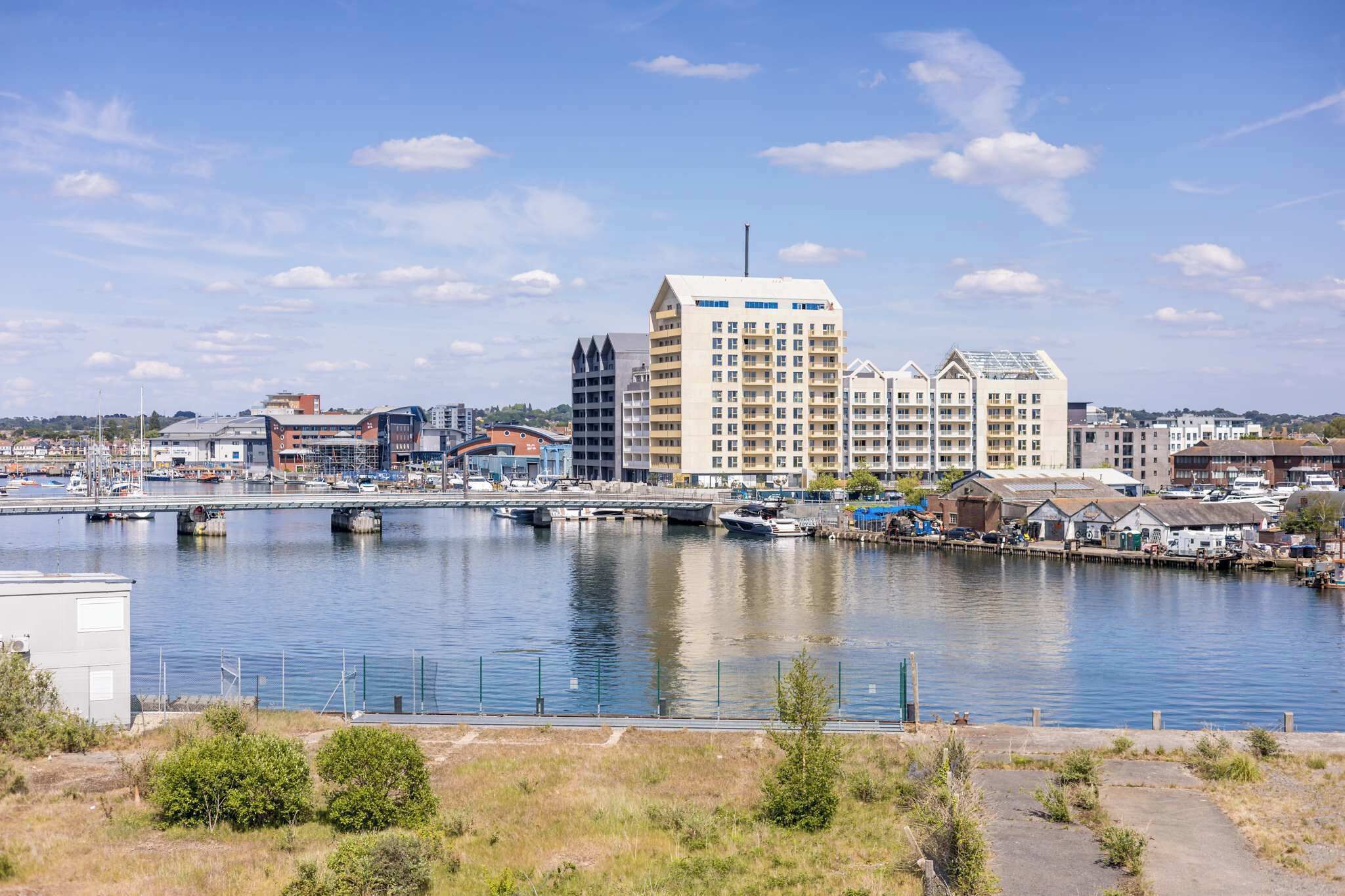 A waterfront cityscape with modern buildings, a marina, and a bridge.
