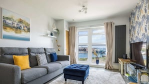 A modern living room with a grey sofa, a blue ottoman, and a view of a river. - Sun-Soaked - Charming Views Overlooking the Harbour (Poole)
