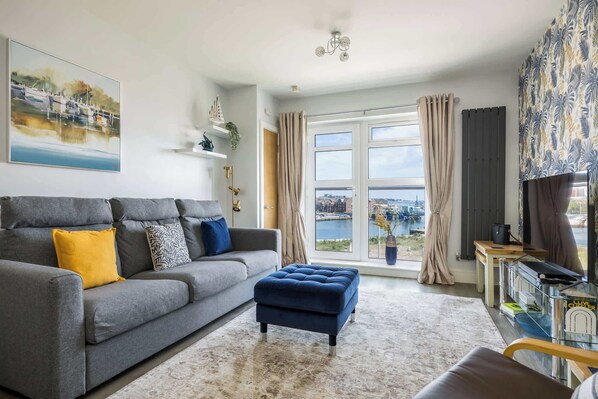 Un salon moderne avec un canapé gris, un pouf bleu et une vue sur une rivière.