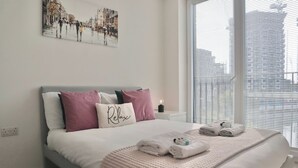 A neatly made bed with a white comforter and pink pillows, a bedside table with a candle, and a window with blinds. - Stylish Riverside Balcony Flat Near Canary Wharf (Greater London)