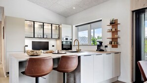 Una cocina moderna con una isla central, dos taburetes de bar, un televisor integrado y una ventana con vistas a zonas verdes.