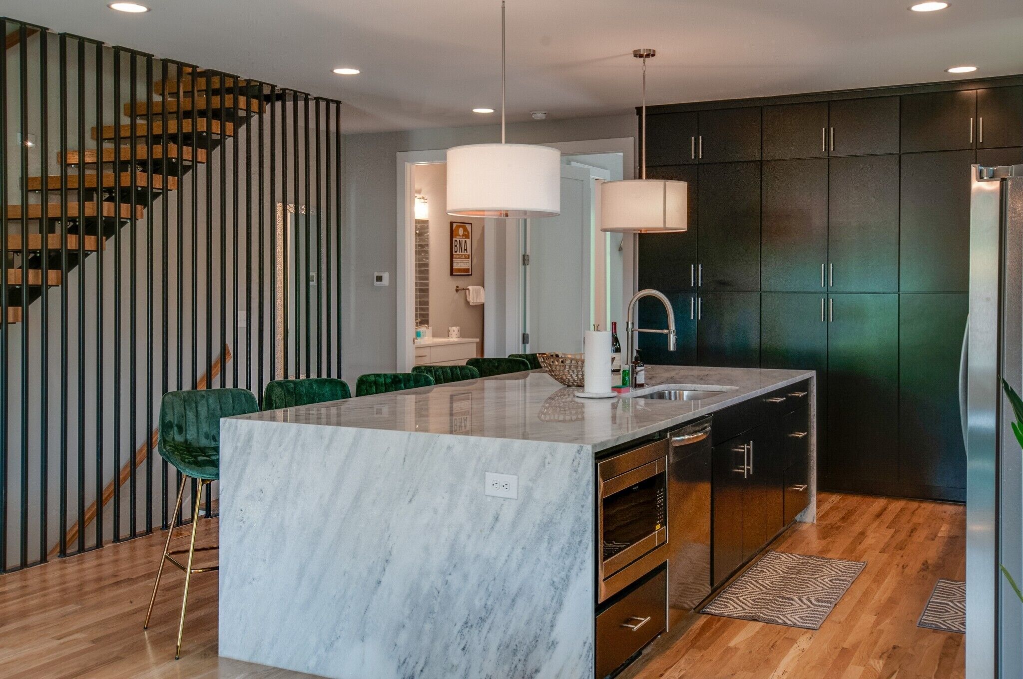 A modern kitchen with a marble island, dark cabinets, and a wooden staircase.