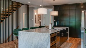 A modern kitchen with a marble island, dark cabinets, and a wooden staircase. - Luxury 10-Bed Home w/ HUGE Rooftop & Game Room! (Nashville)