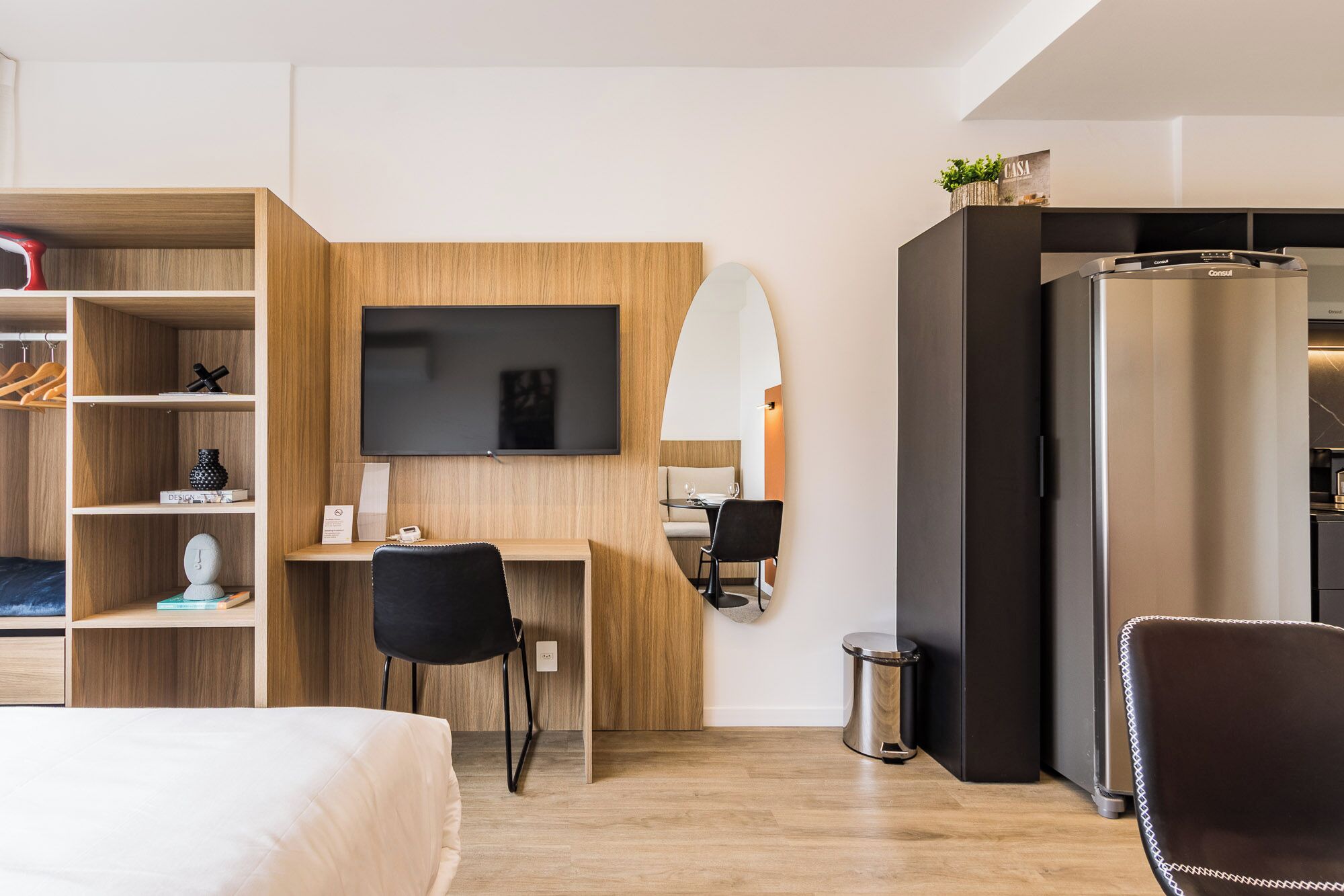 A modern hotel room with a wooden desk, a flat-screen TV, a bed, a dining area, and a minibar.