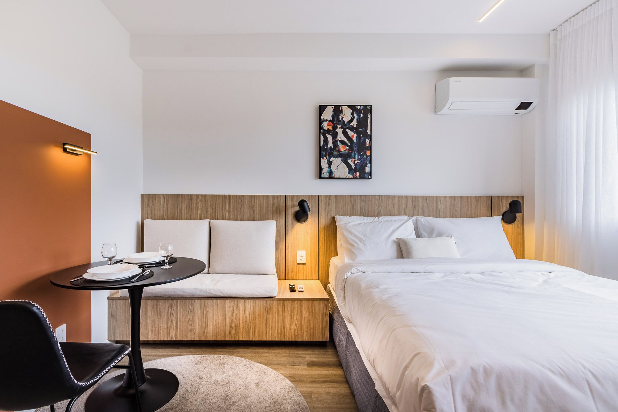 A hotel room with a bed, a small table with a chair, a wall-mounted air conditioner, and a painting on the wall.