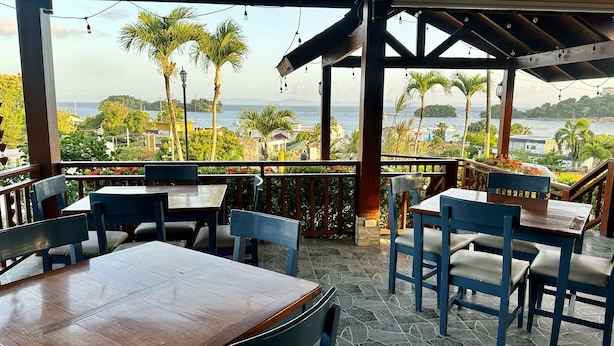 A covered outdoor seating area with tables and chairs, overlooking a scenic view of the sea and palm trees. - Hotel & Restaurant Chino (Samaná)
