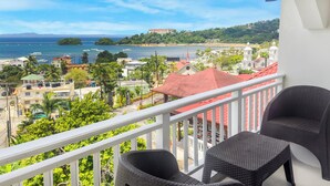 A balcony with wicker chairs overlooking a coastal town with a bridge, buildings, and a hill with a large structure. - Hotel & Restaurant Chino (Samaná)