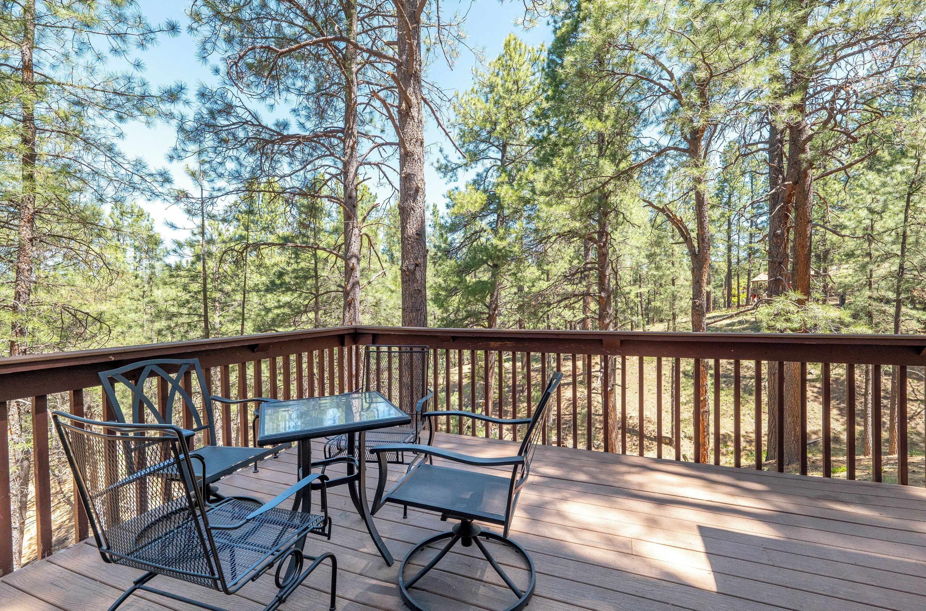 Une terrasse en bois, avec des chaises et une table en métal, entourée de grands pins.