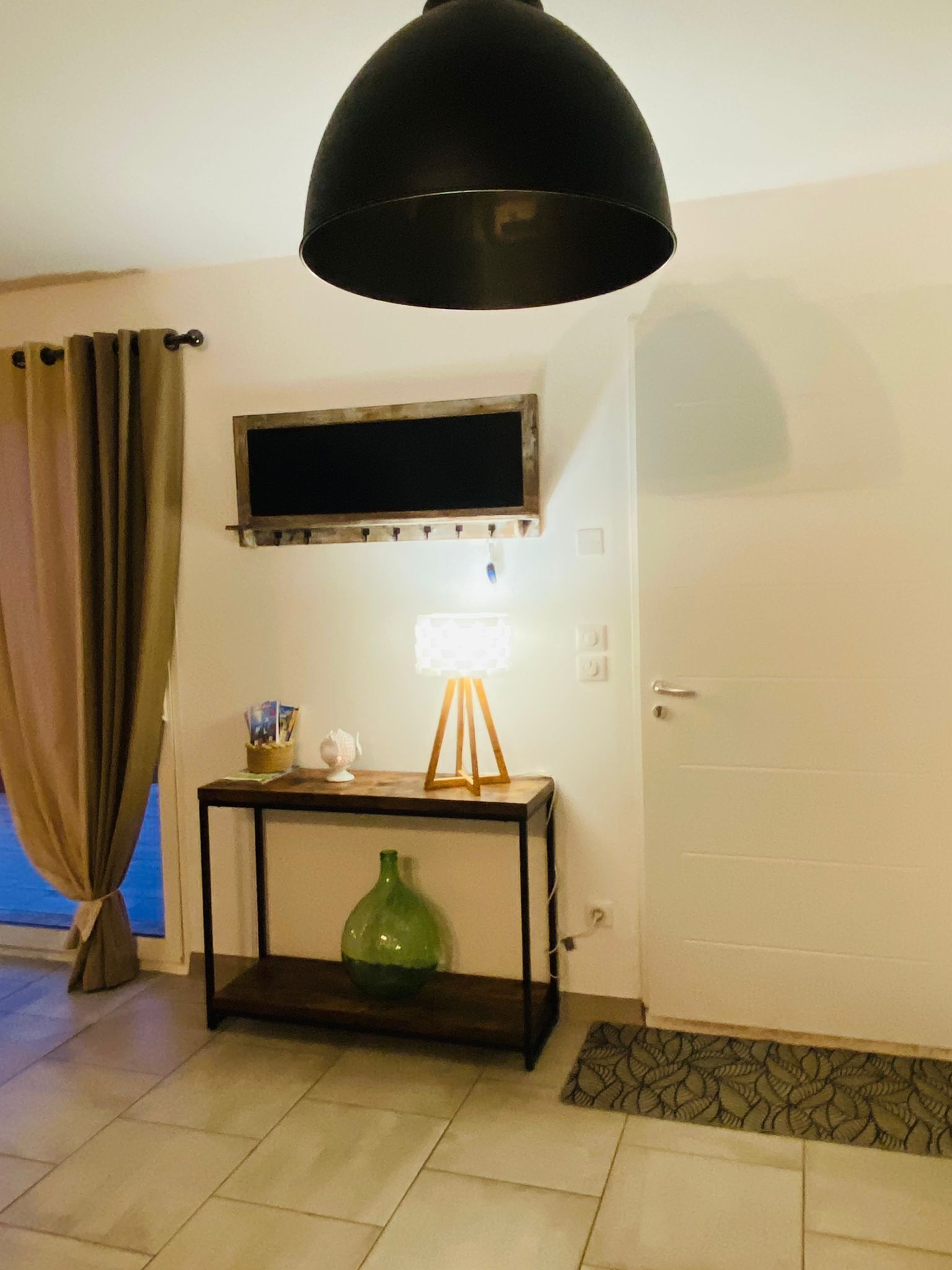 Ein Zimmer mit einer schwarzen Pendelleuchte, einem kleinen Tisch mit einer grünen Vase, einem an der Wand montierten Fernseher und einer Tür mit einem gemusterten Fußmatten.