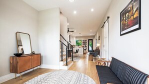 Ein geräumiges Wohnzimmer mit einer schwarzen Ledersofa, einem Holztisch und einem gemusterten Teppich. Ein großer Spiegel befindet sich an der Wand, und ein gerahmtes Kunstwerk ist über dem Sofa angebracht.