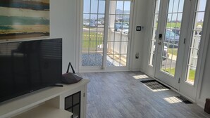 Sala de estar con televisor de pantalla plana, un cuadro y vista a un estacionamiento a través de amplias puertas de vidrio.