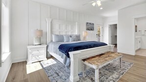 Une chambre à coucher comprenant un lit, des tables de chevet, un banc et un ventilateur de plafond, avec vue sur la salle de bain adjacente.