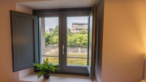 Une chambre avec une fenêtre donnant sur une rivière et des bâtiments, un lit avec une tête de lit blanche, et une plante en pot sur le rebord de la fenêtre.