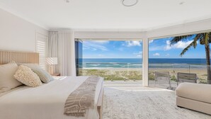 Ein Schlafzimmer mit einem großen Bett, einem Nachttisch, einer Lampe und Blick auf Strand und Meer durch eine Schiebetür.