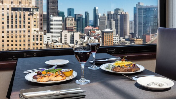 A dining table with plates of food, two wine glasses, and cutlery, set against a cityscape view. - Marines Memorial Club & Hotel  (San Francisco)