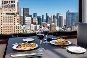 Une table à manger dressée avec des assiettes de nourriture, deux verres à vin et des couverts, le tout mis en valeur par une vue sur le paysage urbain.