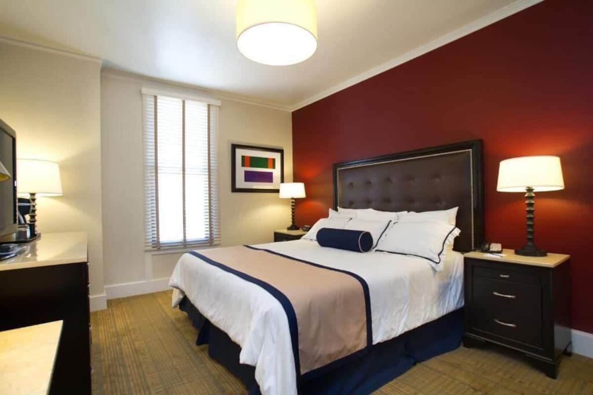 Quarto de hotel com uma cama grande, duas mesas de cabeceira com luminárias, uma janela com persianas e um quadro na parede.