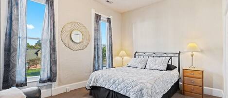 A bedroom with a bed, two bedside tables, a ceiling fan, and a view of the outdoors.