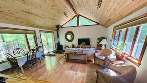 Un salon spacieux doté d’un plancher en bois, un coin repas avec une table et des chaises, un canapé et un téléviseur. De grandes fenêtres laissent entrer la lumière naturelle et offrent une vue sur la verdure.
