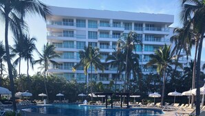 A swimming pool with lounge chairs and umbrellas, surrounded by palm trees and a multi-story building. - Hotel Sea Garden, Nuevo Vallarta, Mexico. Sleeps 4 adults & 2 children. (Nuevo Vallarta)