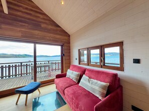 A room with a red sofa, a blue chair, and a glass table. There are large windows with wooden frames, and a view of a body of water and hills. - Rare petit chalet sur le lac, barque à rames, clim (Aiguebelette-le-Lac)