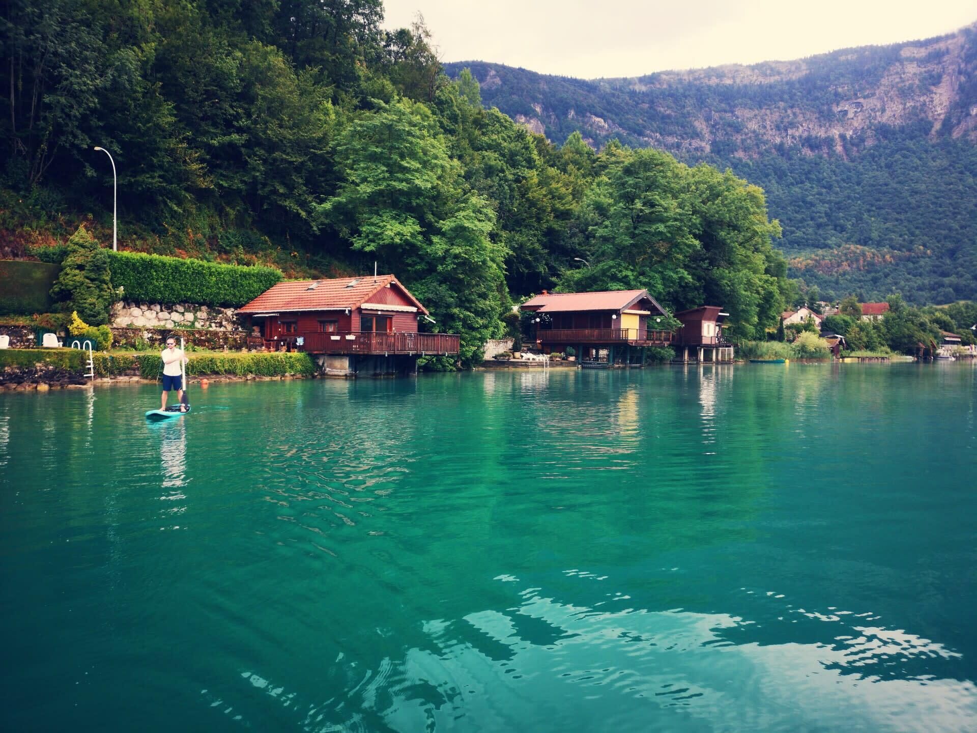 A serene lakeside setting with a house on stilts, surrounded by lush greenery and mountains in the background.