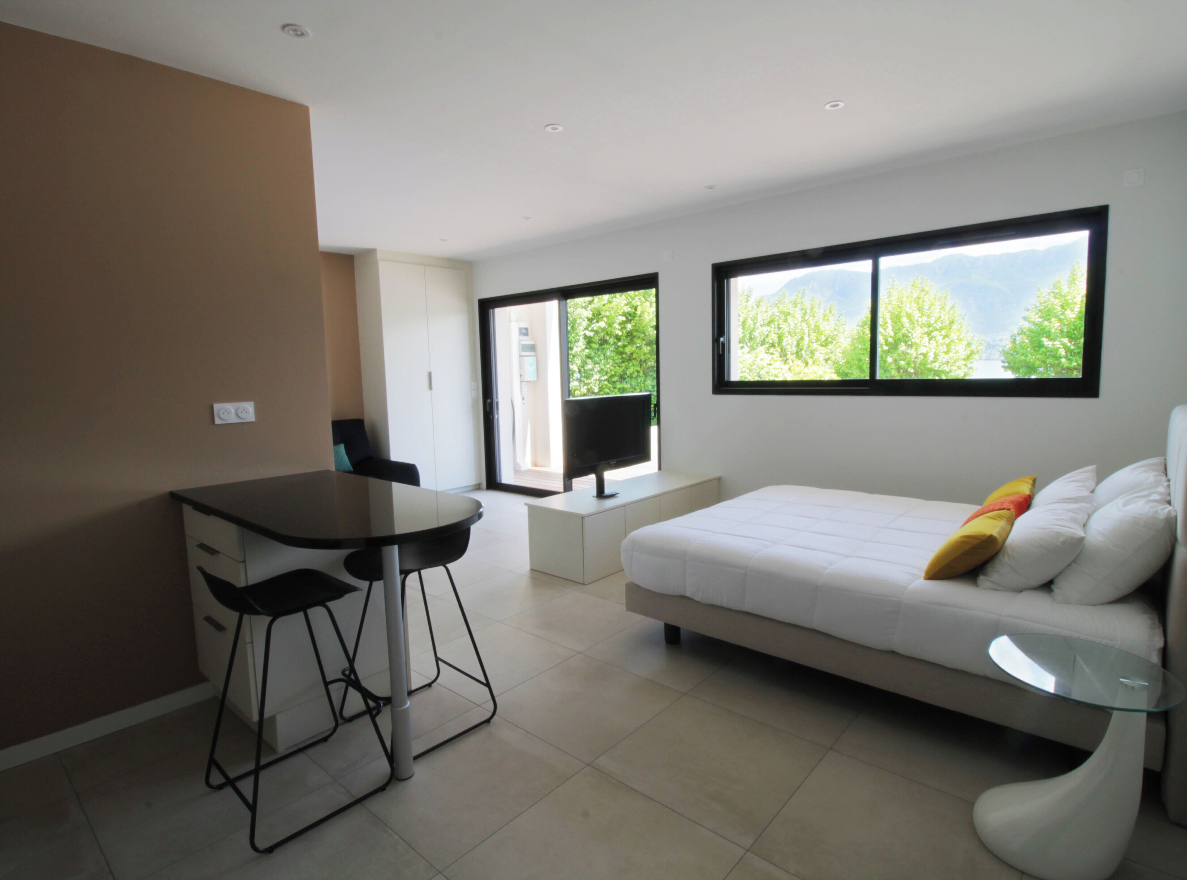 A modern bedroom with a bed, a glass coffee table, a desk with two chairs, and a TV.