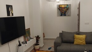 Una sala de estar con una televisión de pantalla plana, un sofá gris con un cojín amarillo, un cuadro en la pared y una planta en maceta.