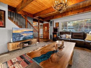 A spacious living room with a wooden coffee table, a large flat-screen TV, a leather sofa, and a staircase with wooden railings. - Birdhouse in Moonridge (Big Bear City)