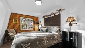 Un dormitorio con una cama grande, cabecera de madera, ventana con vistas a árboles, una silla con una manta y una mesita de noche con lámpara.