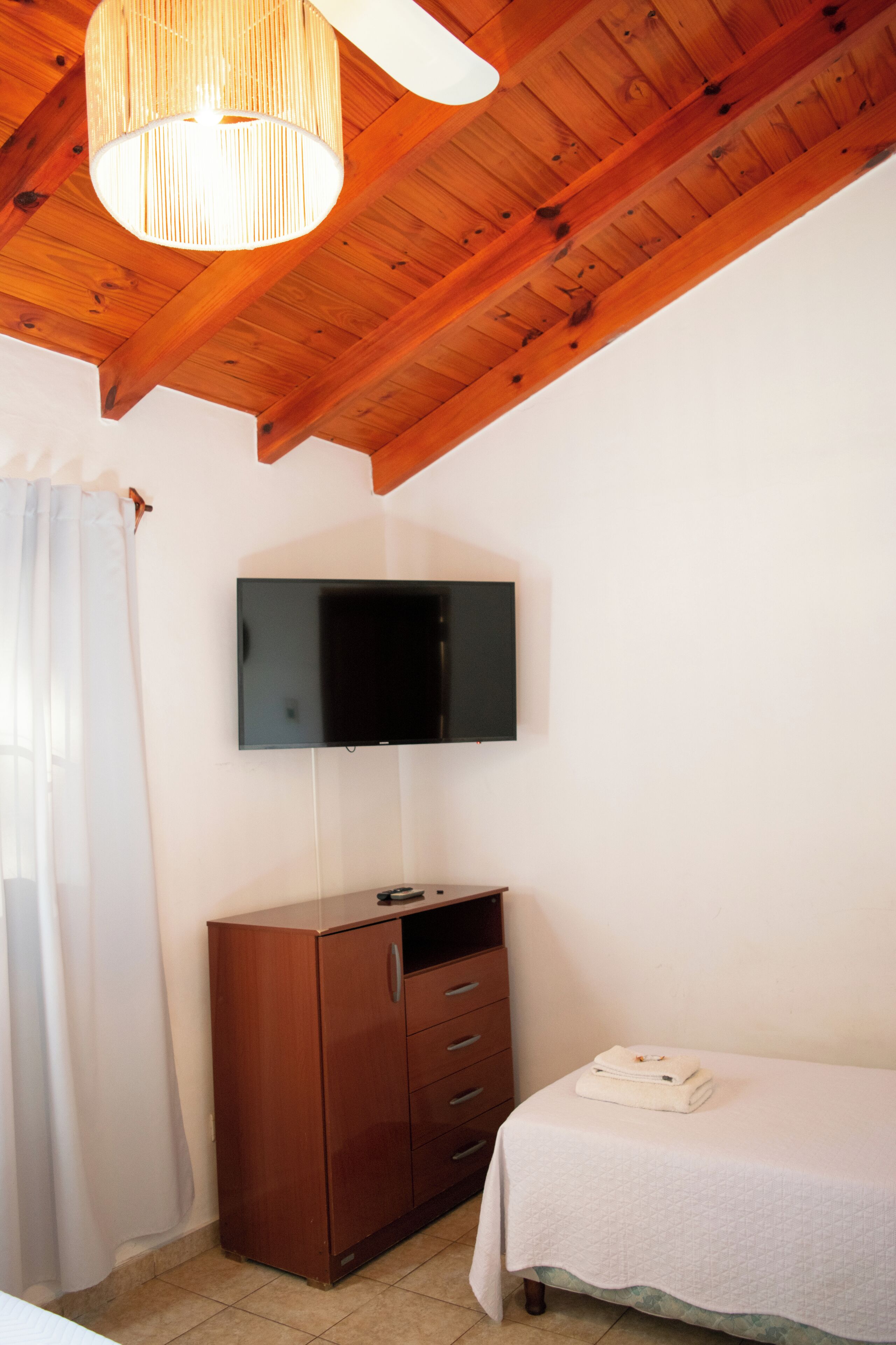 A bedroom with a wooden ceiling, a flat-screen TV mounted on the wall, a wooden dresser, and a bed with a white bedspread.