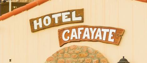 A hotel named 'CAFAYATE' with a stone facade, a metal gate, and signs indicating 'PILETA DESAYUNO' and 'Wi-Fi' availability.