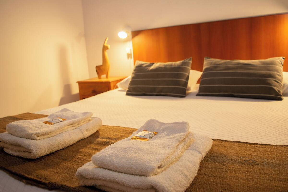 A neatly made bed with two pillows, a bedside table with a lamp, and two folded towels on a brown mat.