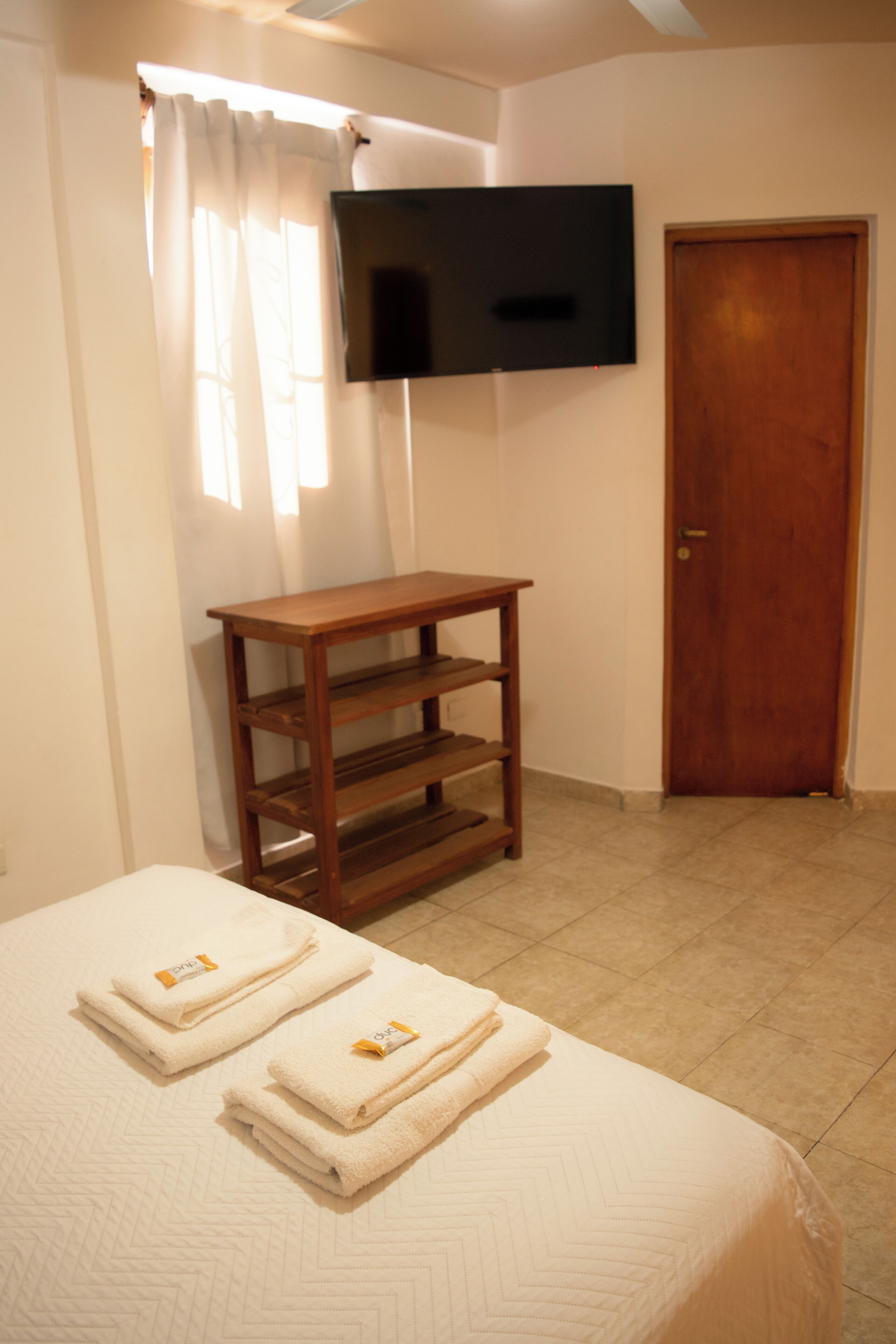 Una habitación con cama, una mesa de madera, televisor montado en la pared y una puerta.