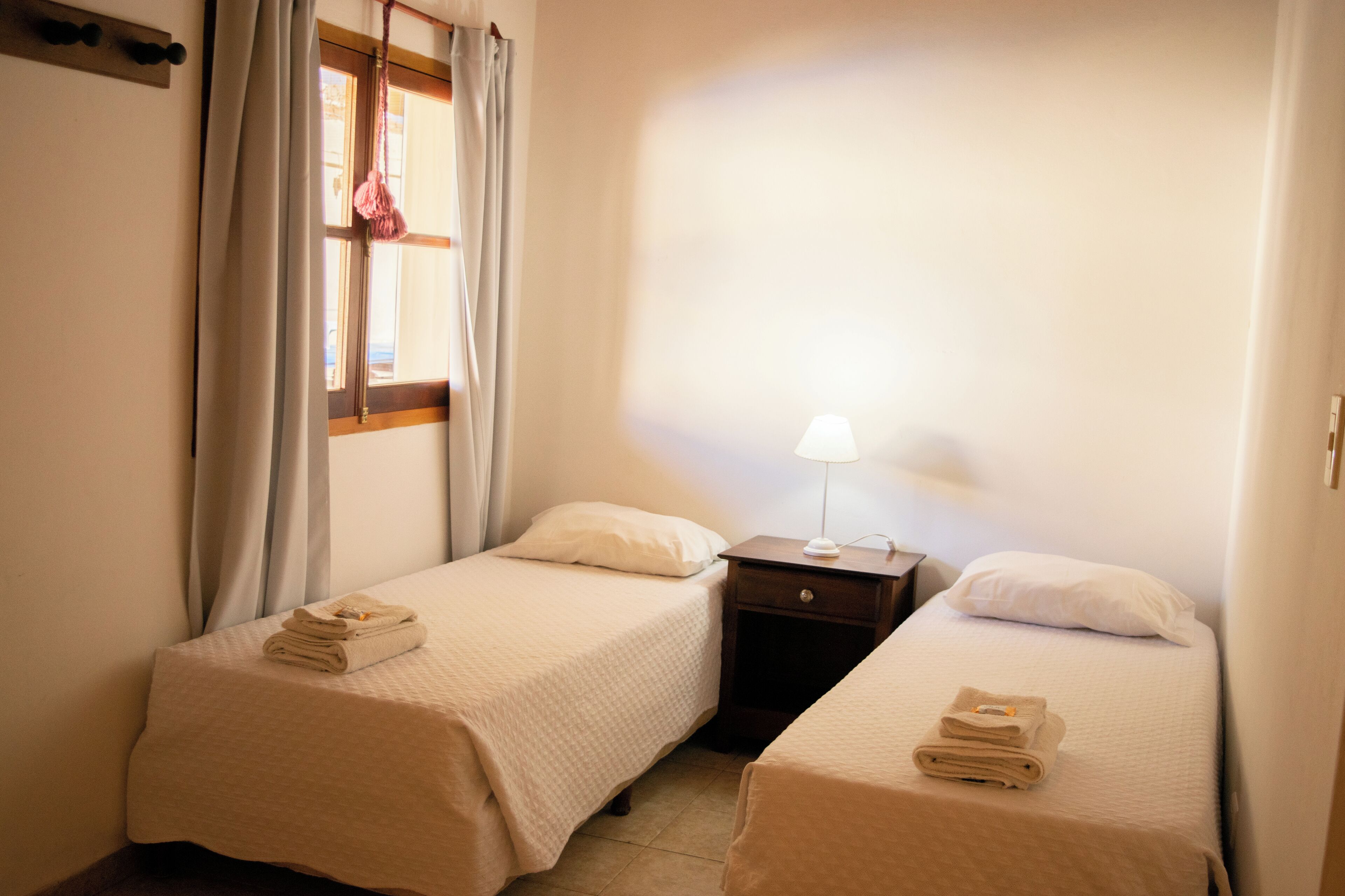 A hotel room with two single beds, each with a neatly folded towel, a bedside table with a lamp, and a window with curtains.
