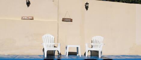 A poolside area with two white chairs and a small table, a sign indicating pool hours, and two wall-mounted lights.