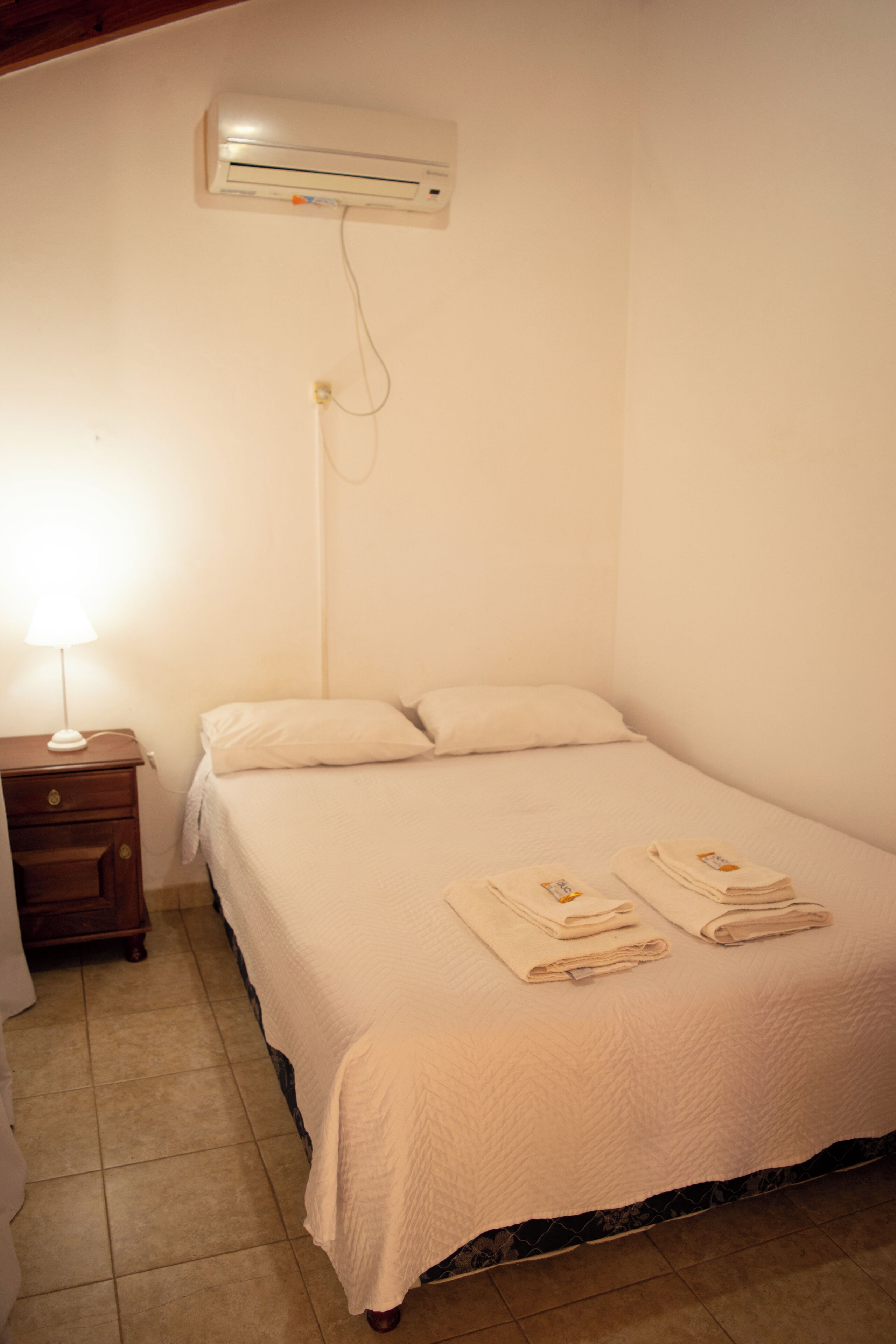 A single bed with a white comforter and two folded towels, a bedside table with a lamp, and an air conditioning unit mounted on the wall.