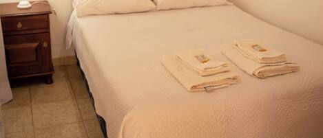 A single bed with a white comforter and two folded towels, a bedside table with a lamp, and an air conditioning unit mounted on the wall.
