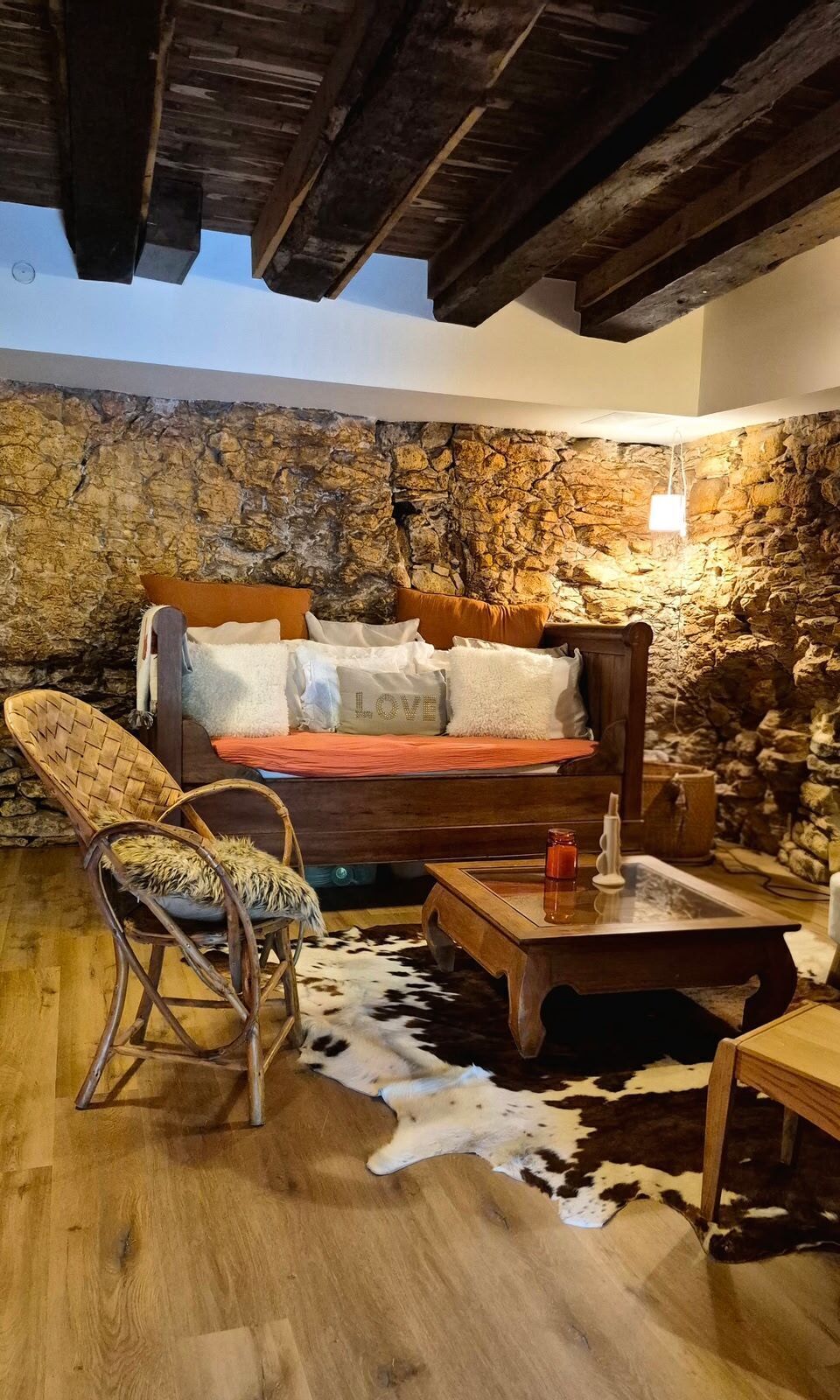 A rustic room with a stone wall, wooden ceiling beams, a bed, a woven chair, a wooden table, and a cowhide rug.
