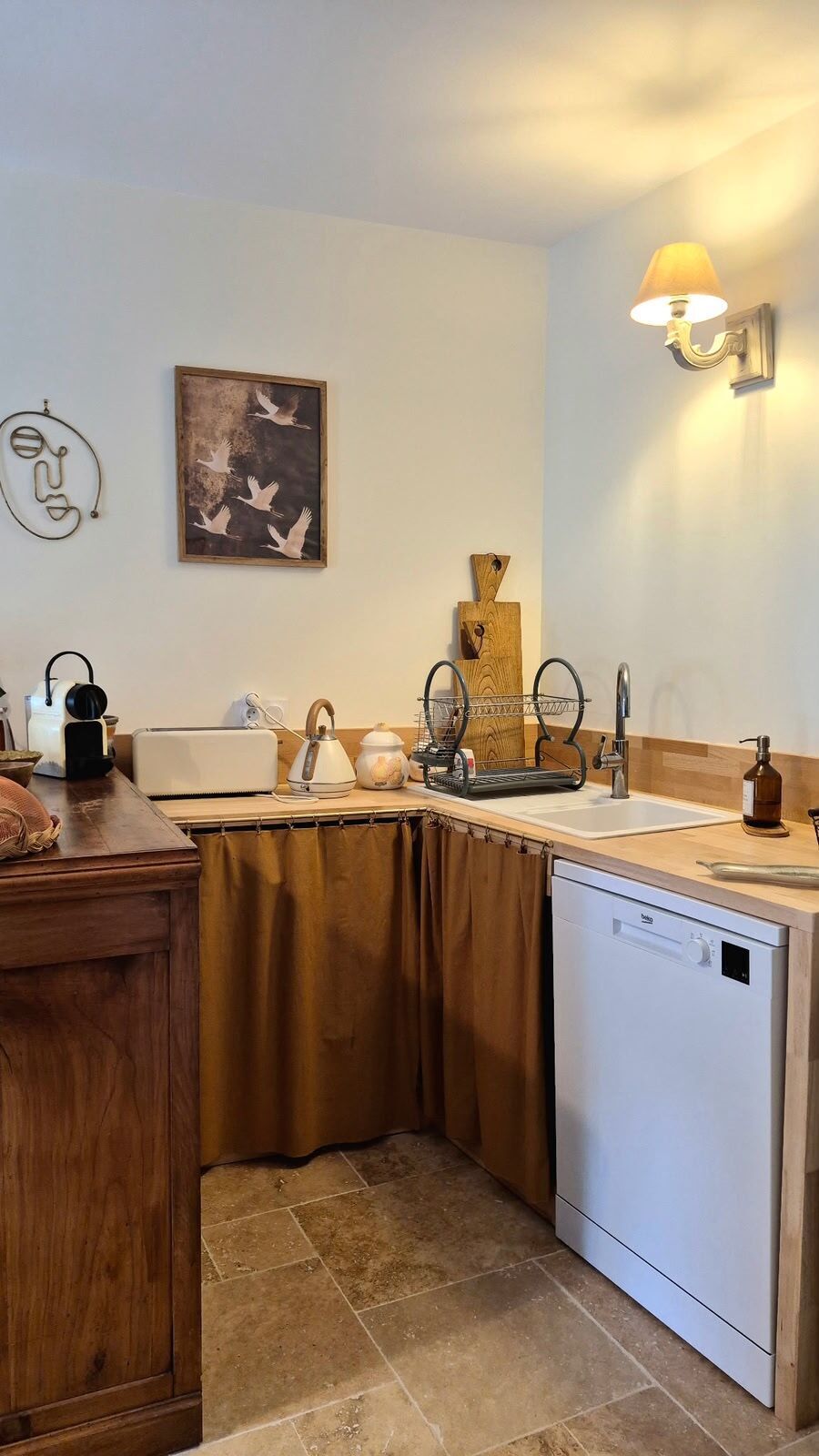A kitchen with a dishwasher, a sink, a countertop with a kettle, a toaster, and a decorative wooden piece.