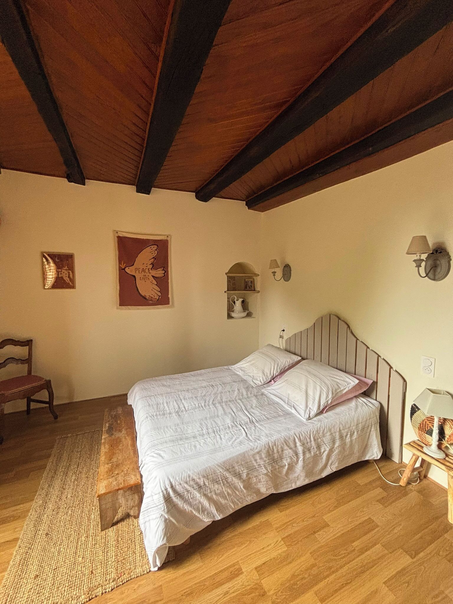 A bedroom with a wooden ceiling, a bed with white bedding, a wooden floor, a chair, and wall-mounted lamps.
