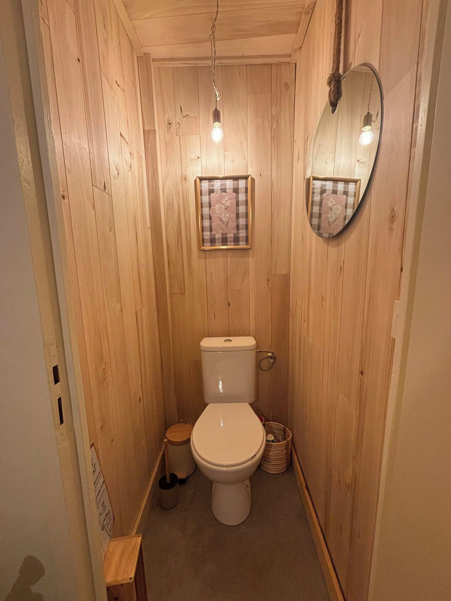 A small, wooden-paneled bathroom with a toilet, a framed floral artwork, a hanging light bulb, and a mirror on the wall.