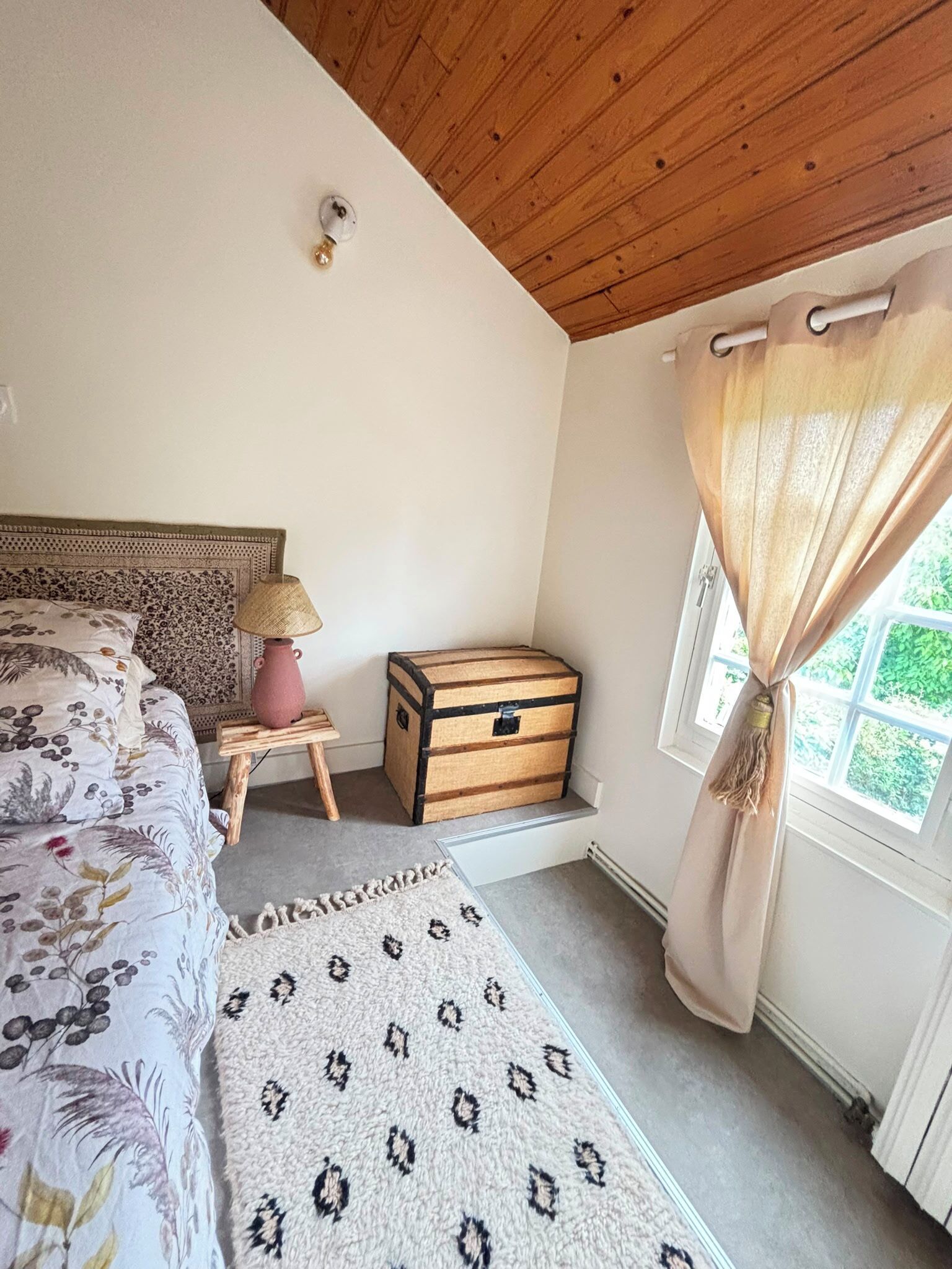 A bedroom with a bed, a small table with a lamp, a chest of drawers, a patterned rug, and a window with curtains.