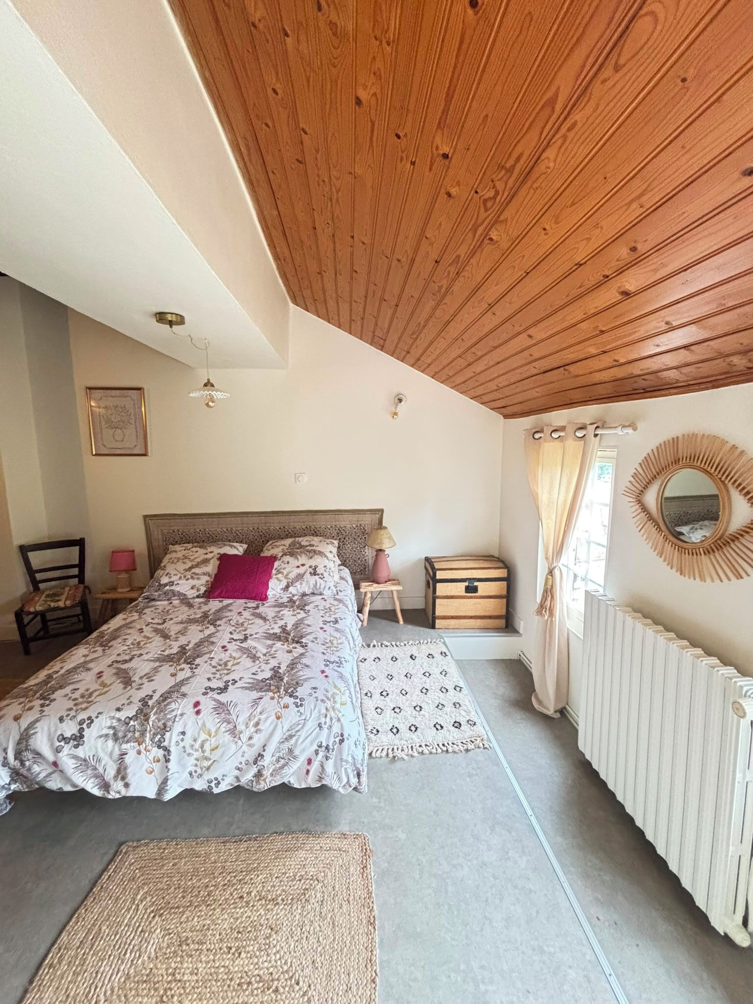 Un dormitorio con techo de madera, una cama con ropa de cama floral, una mesita de noche con lámpara, una silla, una ventana con cortinas y un radiador.