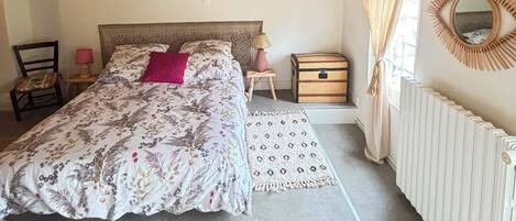 Un dormitorio con techo de madera, una cama con ropa de cama floral, una mesita de noche con lámpara, una silla, una ventana con cortinas y un radiador.