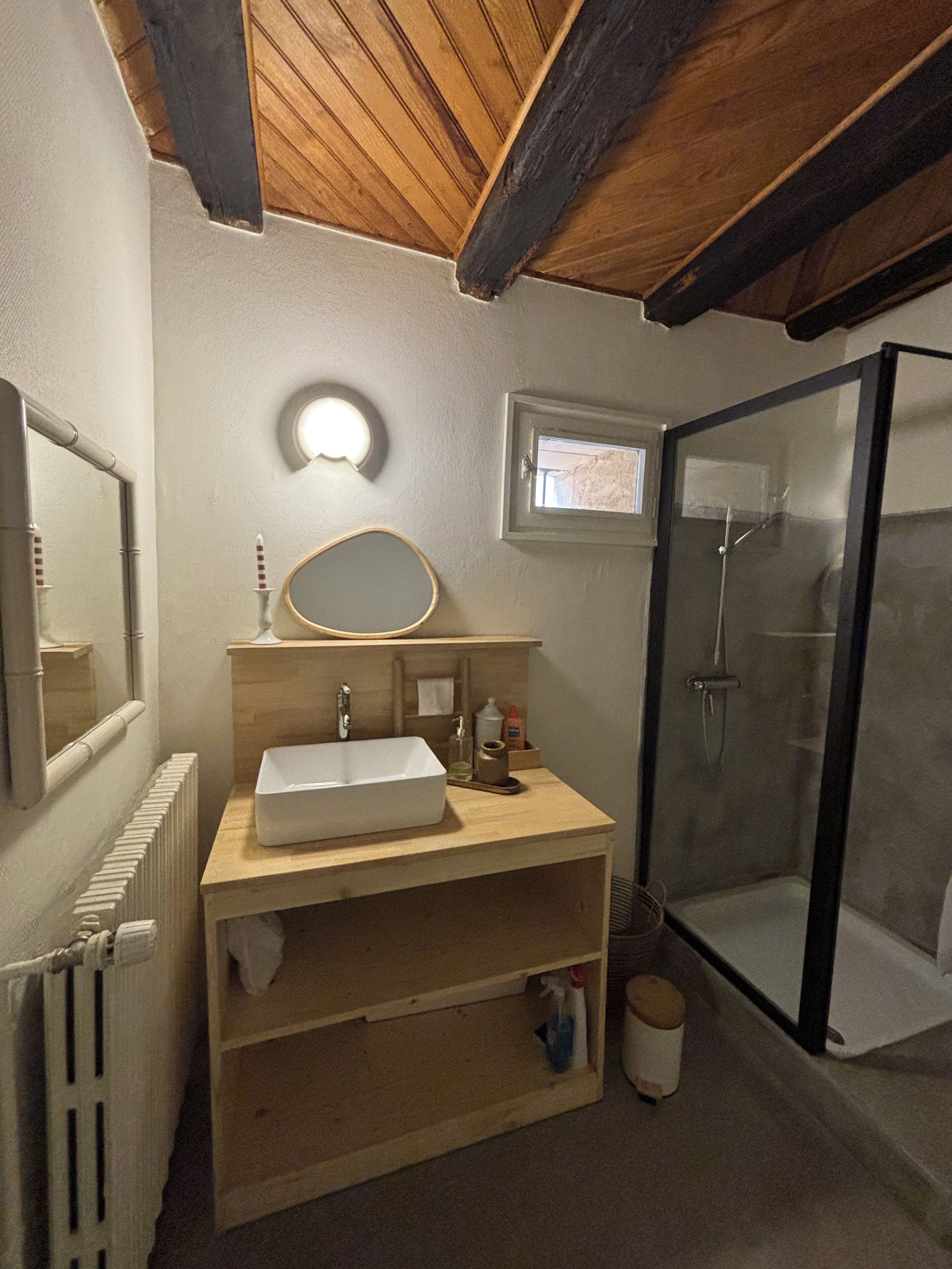 A bathroom with a wooden vanity, a white sink, a mirror, a small window, and a glass shower enclosure.