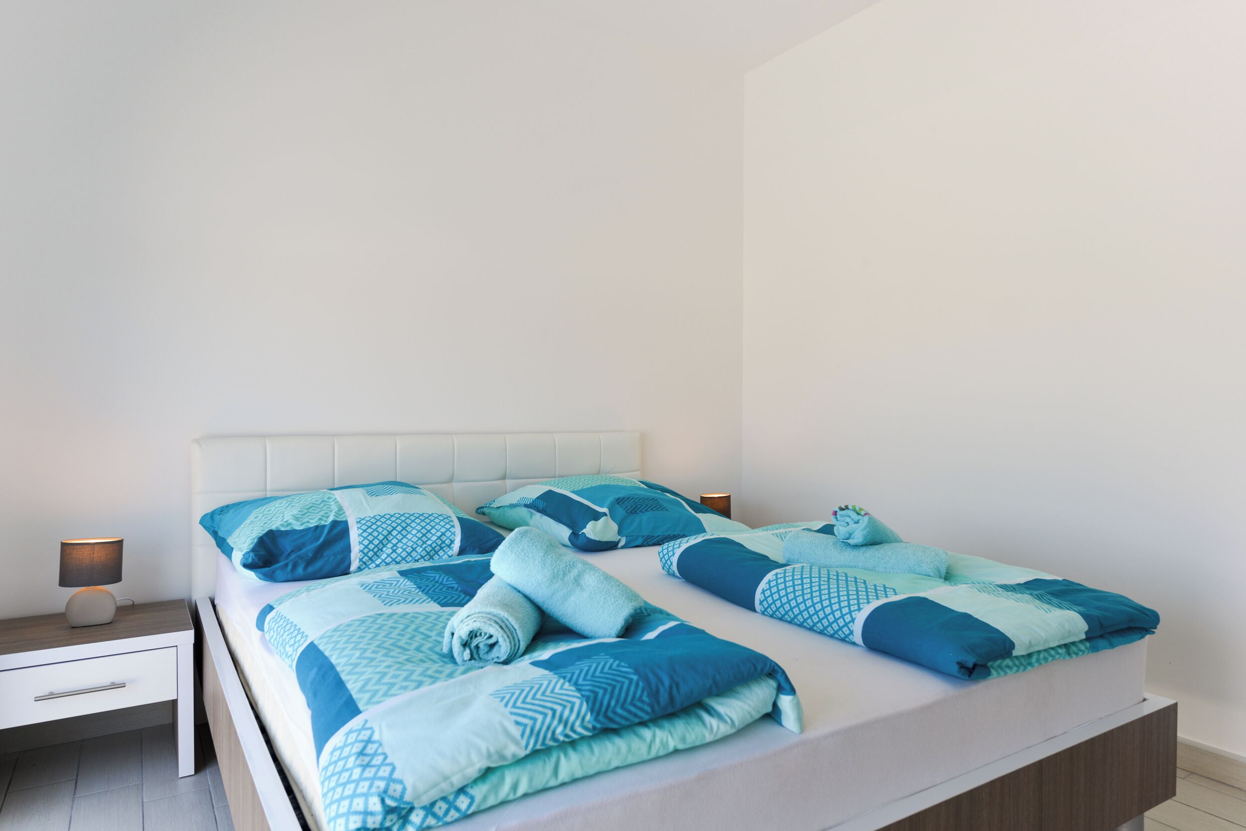 Una cama bien hecha con ropa de cama azul y blanca, una mesita de noche con una lámpara y una mesita auxiliar de madera.